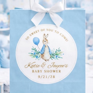 Pode incluir: Uma sacola de presente azul claro para chá de bebê com laço e alças brancas. A sacola apresenta um adesivo circular com um coelho segurando um balão azul e o texto "So Sweet of You to Come!" e "Katie & Jaycee's BABY SHOWER 9/21/28".