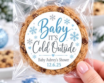 May include: A chocolate chip cookie in a clear plastic bag with a round white sticker featuring blue snowflakes and the text Baby It's Cold Outside, Baby Aubrey's Shower, and the date 12.6.25 printed in blue and gray ink on the front surface.