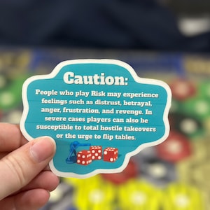 Op de afbeelding: Een blauwe en witte sticker met de tekst "Let op: Mensen die Risk spelen kunnen gevoelens ervaren zoals wantrouwen, verraad, woede, frustratie en wraak. In ernstige gevallen kunnen spelers ook vatbaar zijn voor totale vijandige overnames of de drang om de tafel om te gooien." De sticker bevat ook een kleine illustratie van een spelstuk en drie dobbelstenen.