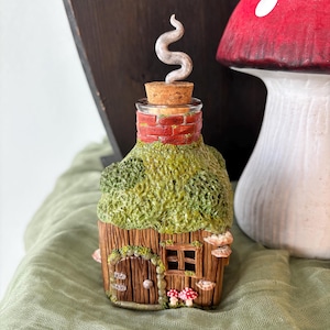 May include: A glass bottle shaped like a small cottage with a brown wood-textured exterior, a green mossy roof, a red brick chimney, and a cork stopper topped with a swirling white smoke sculpture. Two small red and white mushrooms sit by the door.