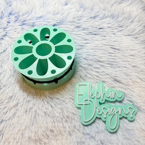 May include: A mint green, flower-shaped container with a black band around the middle. The container has several holes and a central opening. Next to it is a mint green sign with the text "Elephin Designs" in white.