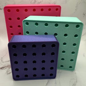 May include: Three square storage boxes with holes in the top. The boxes are stacked on top of each other. The top box is pink, the middle box is light green, and the bottom box is purple.