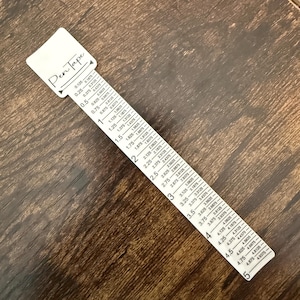 May include: A white Pen Tape ruler with black markings and measurements in inches. The ruler has the text "Pen Tape" printed on the top and is placed on a brown wooden surface.