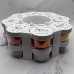 May include: A white plastic glitter organizer filled with small jars of micro fine glitter in various colors. The jars have gray lids and labels with text. The glitter colors include red, orange, silver, and yellow. The text on the labels includes "Crimson" and "A Lil' Rusty".