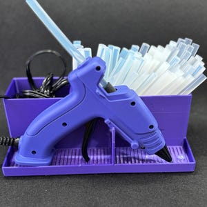 May include: A purple glue gun with a black trigger and nozzle, resting in a matching purple plastic organizer stand. The stand contains a bundle of translucent white glue sticks and a black power cord coiled in the back compartment.