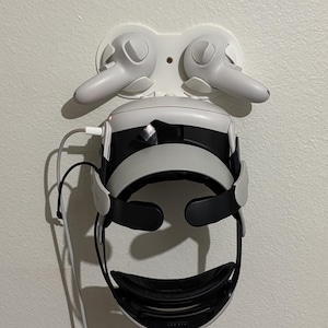 May include: A white wall-mounted storage dock holding a virtual reality headset and two matching white controllers. The headset features a black face interface and an adjustable head strap, with a charging cable plugged into the side of the device.