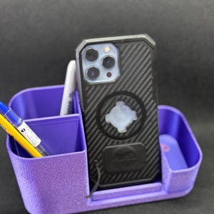 May include: A purple desk organizer with a textured finish featuring multiple compartments. A black smartphone with a carbon fiber pattern case and a circular magnetic mount is positioned in the center slot, alongside a yellow pen and a white marker.
