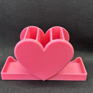 May include: A bright pink desk organizer featuring a prominent heart-shaped front panel. The unit includes two vertical compartments for holding pens or stationery and a wide, rectangular base tray for storing small office supplies or accessories.
