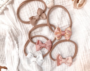 May include: Five baby headbands with thin, stretchy brown nylon bands, each featuring a small grosgrain ribbon bow. The bows are in solid beige, solid dusty rose, solid white, solid light pink, and a multi-colored wavy striped pattern.