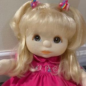 May include: A blonde doll with a pink dress and a white collar. The dress has the word "Love" embroidered on the front. The doll has brown eyes and is wearing two rainbow hair clips.
