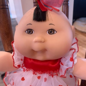 May include: A Cabbage Patch Kids doll with brown eyes and a small black tuft of hair. The doll is dressed in a red and white dress with red heart accents and a matching bow. The doll is seated in a wooden rocking chair.