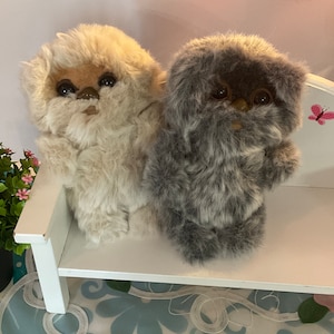 Puede incluir: Dos peluches, uno blanco y otro gris, con caras peludas y ojos grandes. Están sentados en un banco blanco.