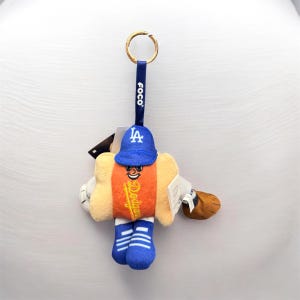 May include: A plush keychain featuring a hot dog character wearing a blue Dodgers baseball cap and uniform. The hot dog has an orange center, a smiling face, and the Dodgers logo. The keychain has a gold ring and a blue strap with the word "FOCO".