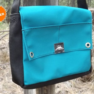 May include: Teal messenger bag with a black base and strap. The bag features a flap with two metal grommets and a small label that reads "OUTROCK". The bag is hanging on a wooden post. A "TOP SELLER" sticker is in the upper left corner.