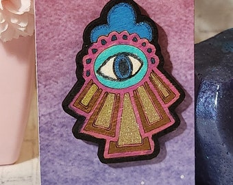 May include: A wooden cutout of a blue, pink, and gold evil eye charm with a white background. The evil eye is a symbol of protection against negative energy.