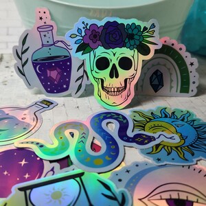 May include: A set of iridescent holographic stickers featuring various mystical and celestial designs, including a skull with a flower crown, a snake, a sun and moon, a bottle with a crescent moon, and a bottle with a crystal.