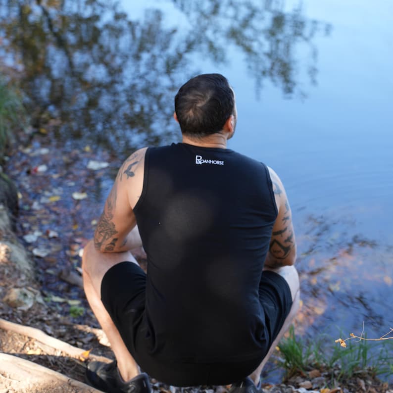 May include: Black tank top with the white text "DANHORSE" on the upper back. The person is wearing black shorts and black shoes. The person is sitting near a body of water.