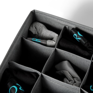 K&ouml;nnte beinhalten: Ein grauer Stoff-Schubladenorganizer mit mehreren F&auml;chern zum Verstauen von Socken oder anderen kleinen Gegenst&auml;nden. Die F&auml;cher sind mit aufgerollten Socken gef&uuml;llt, einige mit dem Buchstaben "R" in Blau bedruckt.