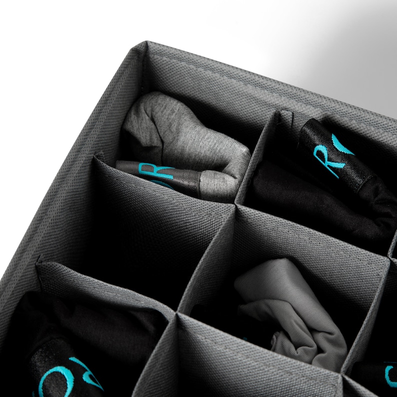 K&ouml;nnte beinhalten: Ein grauer Stoff-Schubladenorganizer mit mehreren F&auml;chern zum Verstauen von Socken oder anderen kleinen Gegenst&auml;nden. Die F&auml;cher sind mit aufgerollten Socken gef&uuml;llt, einige mit dem Buchstaben "R" in Blau bedruckt.