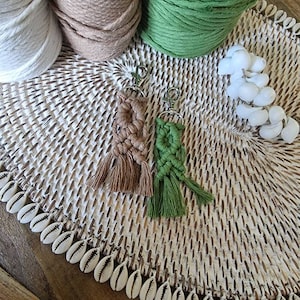 May include: Two macrame keychains with tassels. One is brown and the other is green. They are on a white woven mat with white seashells around the edge.