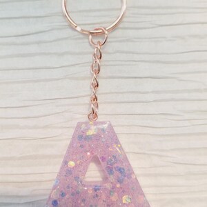 May include: A lavender-colored letter "A" keychain with iridescent glitter embedded in resin. The keychain has a rose gold chain and key ring. The letter is approximately 2 inches tall.