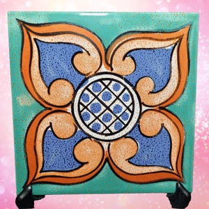 May include: A square ceramic tile featuring a central circular medallion with a blue and white grid pattern. Surrounding the center are four symmetrical orange and blue floral-like shapes set against a speckled turquoise background.