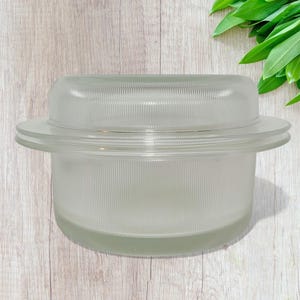 May include: A clear glass bowl with a matching lid. The bowl has a wide, flat rim and vertical ribbed detailing. The lid is also ribbed and fits snugly on top. The glass has a frosted appearance.