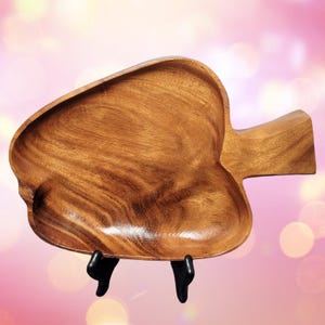 May include: A heart-shaped wooden serving tray with a handle. The tray is made of a light brown wood and is displayed on a black stand. The tray's design is simple and elegant, suitable for serving food or as a decorative piece.
