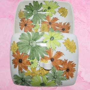 May include: A white ceramic butter dish with a lid, featuring a hand-painted floral pattern in shades of green, yellow, and orange. The rectangular lid includes a small, curved white handle, and the interior of the base shows the same floral design.