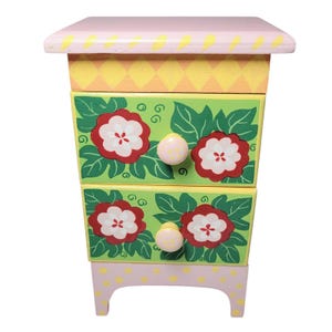 May include: A small, hand-painted wooden cabinet with two drawers. The cabinet features a pink top, yellow and green painted sections, and a pink base with yellow polka dots. The drawers are decorated with red and white floral designs and pink knobs.
