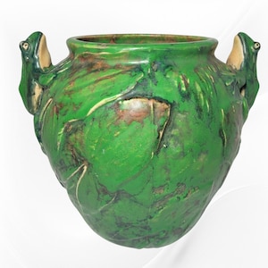 May include: A green ceramic vase with frog handles. The vase has a textured surface with leaf-like patterns and brown accents. The frog handles are light beige with green details. The vase has a wide opening and a rounded body.