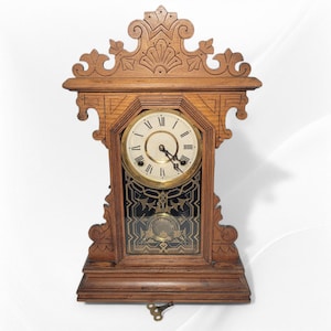 May include: A brown wooden mantel clock featuring an ornate carved top, a circular face with black Roman numerals, and a glass panel below displaying a gold-toned pendulum design. A metal winding key is positioned at the base of the clock.