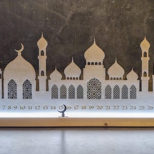May include: A white wooden Ramadan calendar with a crescent moon and a silhouette of a mosque. The calendar has 30 numbered windows, with the numbers 1 through 30 visible. The calendar is lit from behind, creating a warm glow.