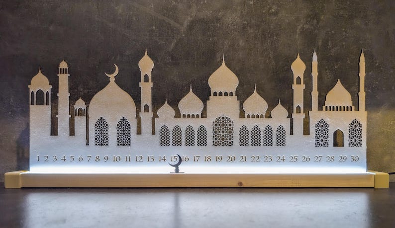 May include: A white wooden Ramadan calendar with a crescent moon and a silhouette of a mosque. The calendar has 30 numbered windows, with the numbers 1 through 30 visible. The calendar is lit from behind, creating a warm glow.