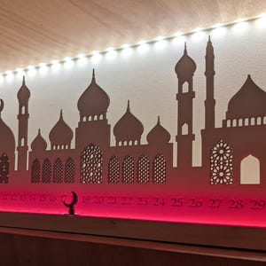 May include: A wooden Ramadan advent calendar with a silhouette of a mosque and a crescent moon. The calendar has numbers 1-30 on the bottom and is lit with red LED lights.