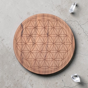 Op de afbeelding: Een ronde houten plank met een lasergeëtst Flower of Life-patroon. De plank is lichtbruin en heeft een donkerbruine rand. Twee heldere bergkristallen liggen in de buurt van de plank op een grijze betonnen ondergrond.