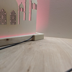 May include: A wooden cutout of a mosque with a red light shining from the base. The cutout is on a wooden base with a black cord.