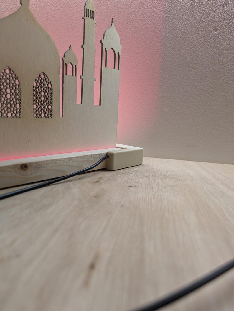 May include: A wooden cutout of a mosque with a red light shining from the base. The cutout is on a wooden base with a black cord.
