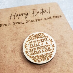 May include: A round wooden ornament with a floral design and the text "Happy Easter" engraved on it. The ornament is on a brown background with the text "Happy Easter! From Greg, Justyna and Ezra" written on it.