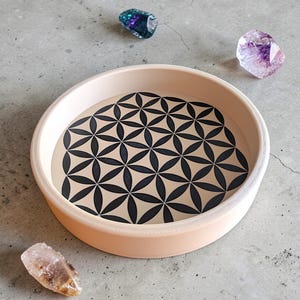 Puede incluir: Una bandeja redonda, de color melocotón claro, con un patrón floral negro y beige en el interior. Tres cristales en tonos morado, azul y transparente están dispersos alrededor de la bandeja sobre una superficie gris. La bandeja es probablemente para fines decorativos.