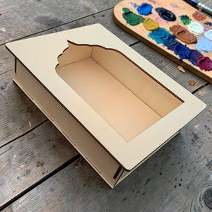 May include: A rectangular light-coloured wooden box with a decorative arched cutout on the top surface. The box features clean, flat edges and a hollow interior, constructed from thin, smooth plywood panels with visible joinery at the corners.