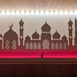 May include: A wooden Ramadan advent calendar with a silhouette of a mosque and crescent moon. The calendar has 28 numbered windows, and the base is lit with red LED lights.