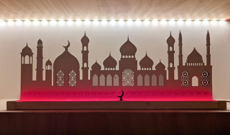May include: A wooden Ramadan advent calendar with a silhouette of a mosque and crescent moon. The calendar has 28 numbered windows, and the base is lit with red LED lights.