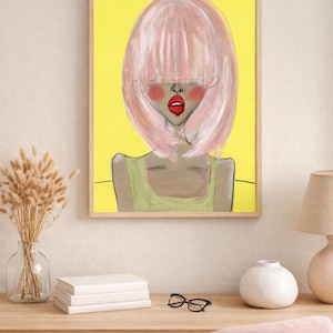 May include: A framed art print featuring a stylized portrait of a person with pink hair, red lips, and rosy cheeks against a yellow background. The person is wearing a green top. The artwork is displayed on a wooden desk with books, glasses, and a vase.