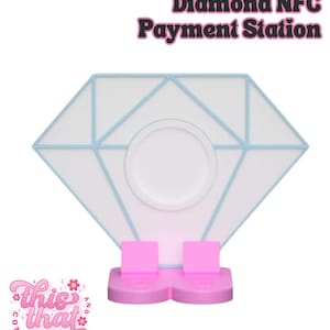 May include: A white diamond-shaped NFC payment station with light blue outlines and a central circular indentation. The base consists of two pink rectangular supports. The text Diamond NFC Payment Station appears above, and a logo for This and That Company is below.