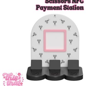 May include: A white NFC payment station with a pink square frame and black base. The station is decorated with small black scissors and the text "Scissors NFC Payment Station". The logo "This That Company" is in the lower left.