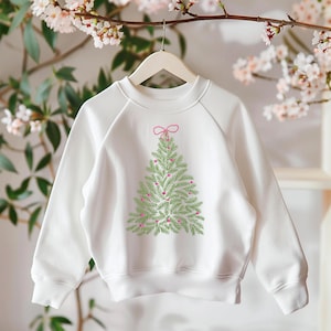 May include: White sweatshirt featuring a green Christmas tree design with pink ornaments and a pink bow at the top. The tree is embroidered on the front of the sweatshirt. The sweatshirt is hanging on a wooden hanger.
