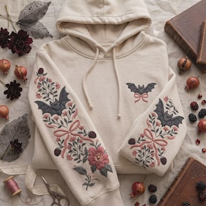 May include: A cream-colored hooded sweatshirt featuring intricate embroidery on both sleeves and the chest. The design includes dark gray bats, pink bows, floral motifs, and dark berries. The hoodie has matching cream-colored drawstrings.