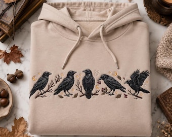 May include: A beige hooded sweatshirt featuring an embroidered design of five black crows perched on branches. The embroidery includes small gold crescent moons, stars, and red berries interspersed between the birds across the chest area.