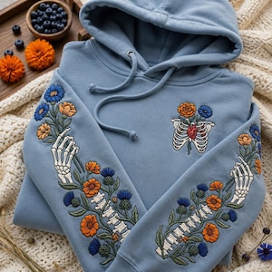 Peut inclure: Un sweat à capuche bleu poussiéreux avec une broderie complexe. Les manches présentent des mains squelettiques tenant des tiges de fleurs orange et bleues. La poitrine montre une cage thoracique avec un cœur rouge et des fleurs.